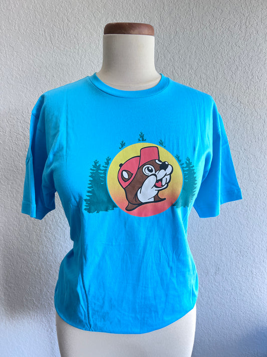 Teal shirt with trees in the middle and a yellow and red circle with Front an image of a beaver in the middle. It's all good in the Woods Blue Buc-ee's Shirt